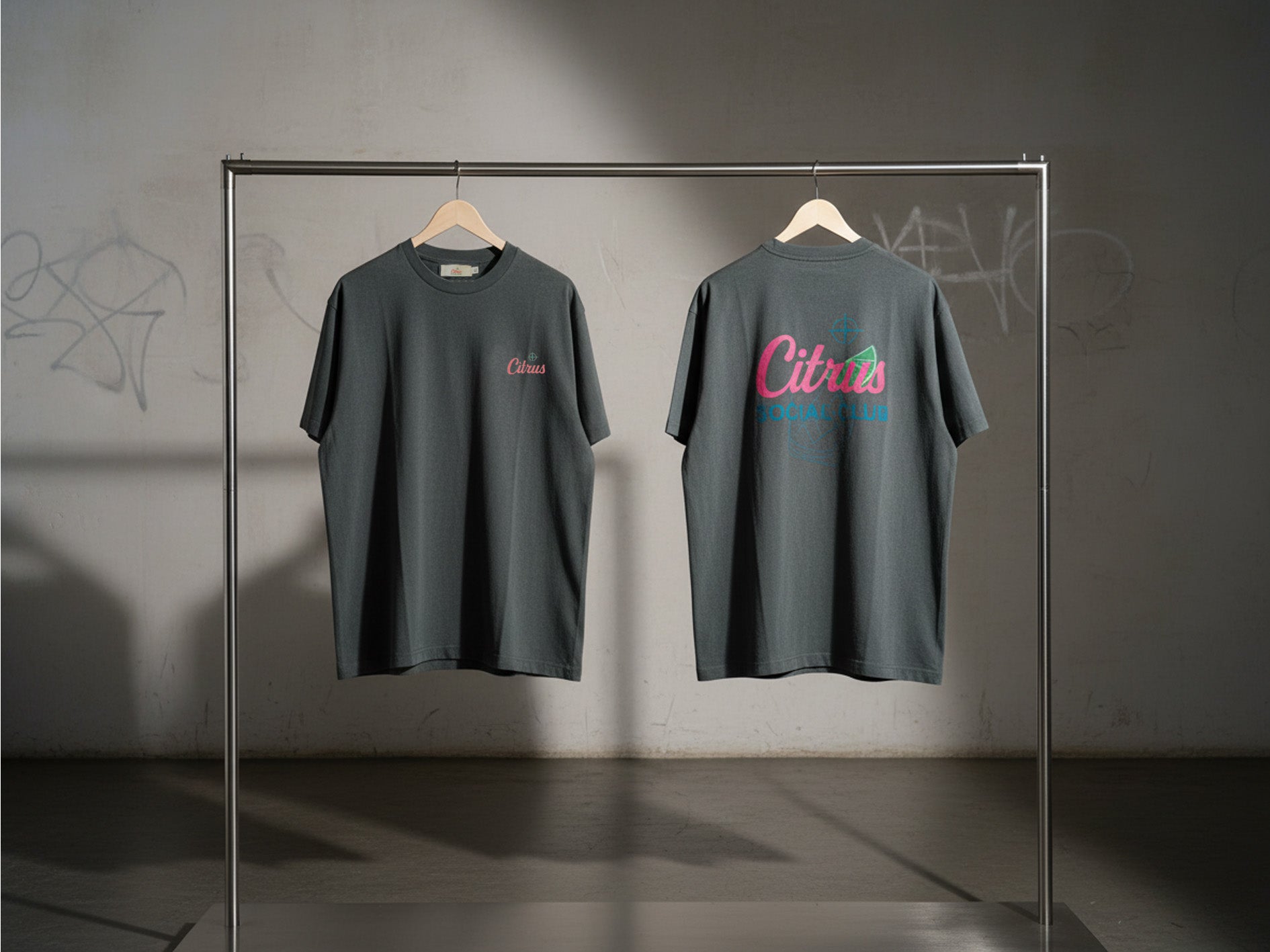 Two dark gray t-shirts with 'Citrus-Social-Club' branding on a rack against a plain wall. One face on pocket positioned logo, one back facing Large Back print Graphic with Margarita style drink and lime.