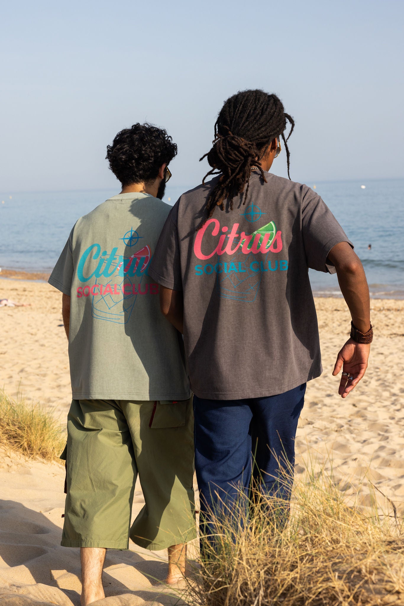 Two people walking on a beach wearing Citrus Social Club t-shirts. Both with picturs of drinks with bright garnish.