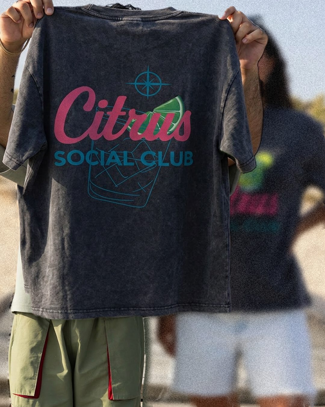 A person outdoors holds up a dark gray washed graphic T-shirt. The shirt has a large front print reading "Citrus Social Club" in pink and teal, with a citrus slice and cocktail glass illustration.