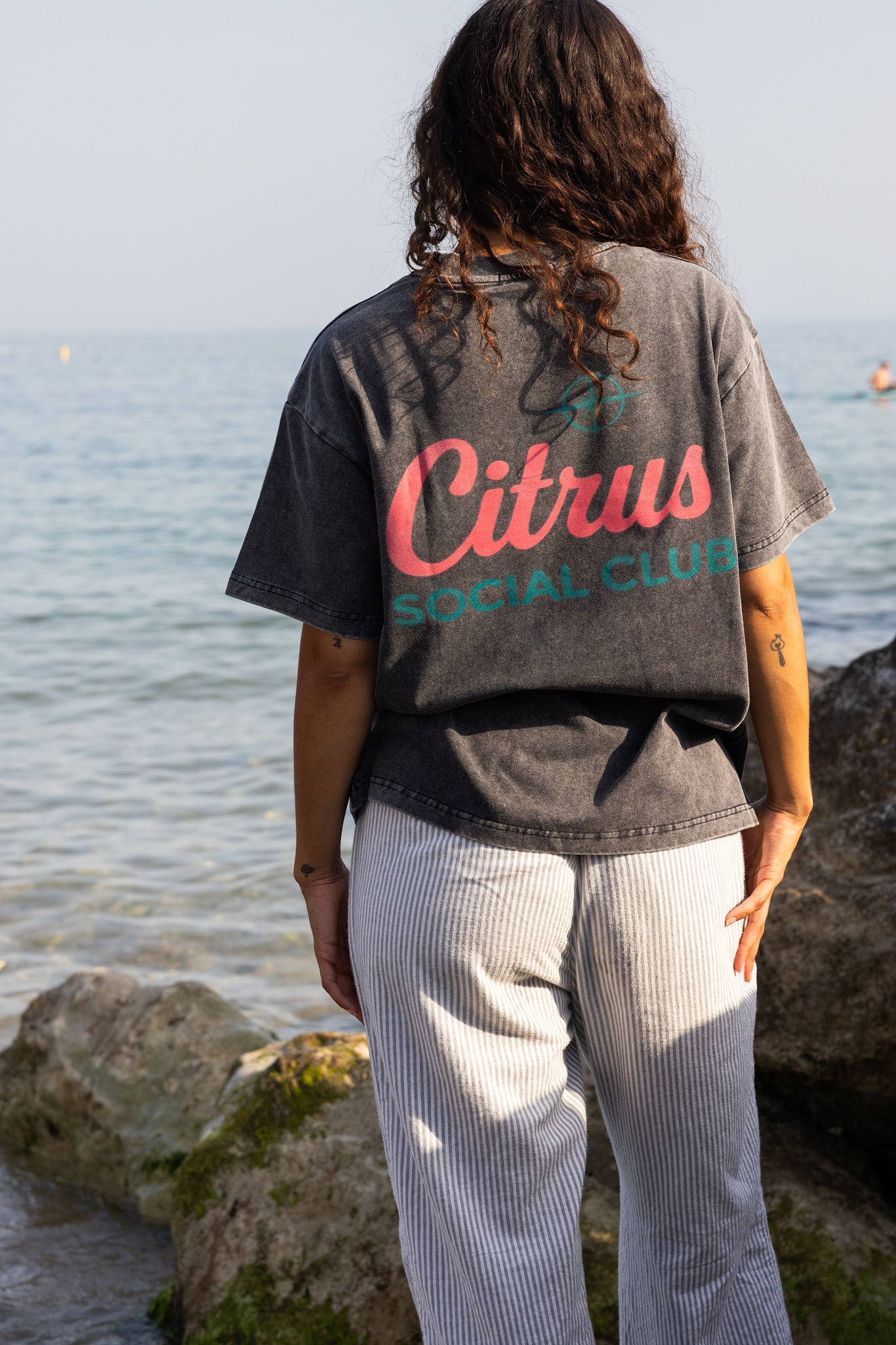 A person standing outdoors by the water wearing a faded dark gray graphic T-shirt with pink and teal lettering that reads 'Citrus Social Club,' paired with light gray pinstriped pants.