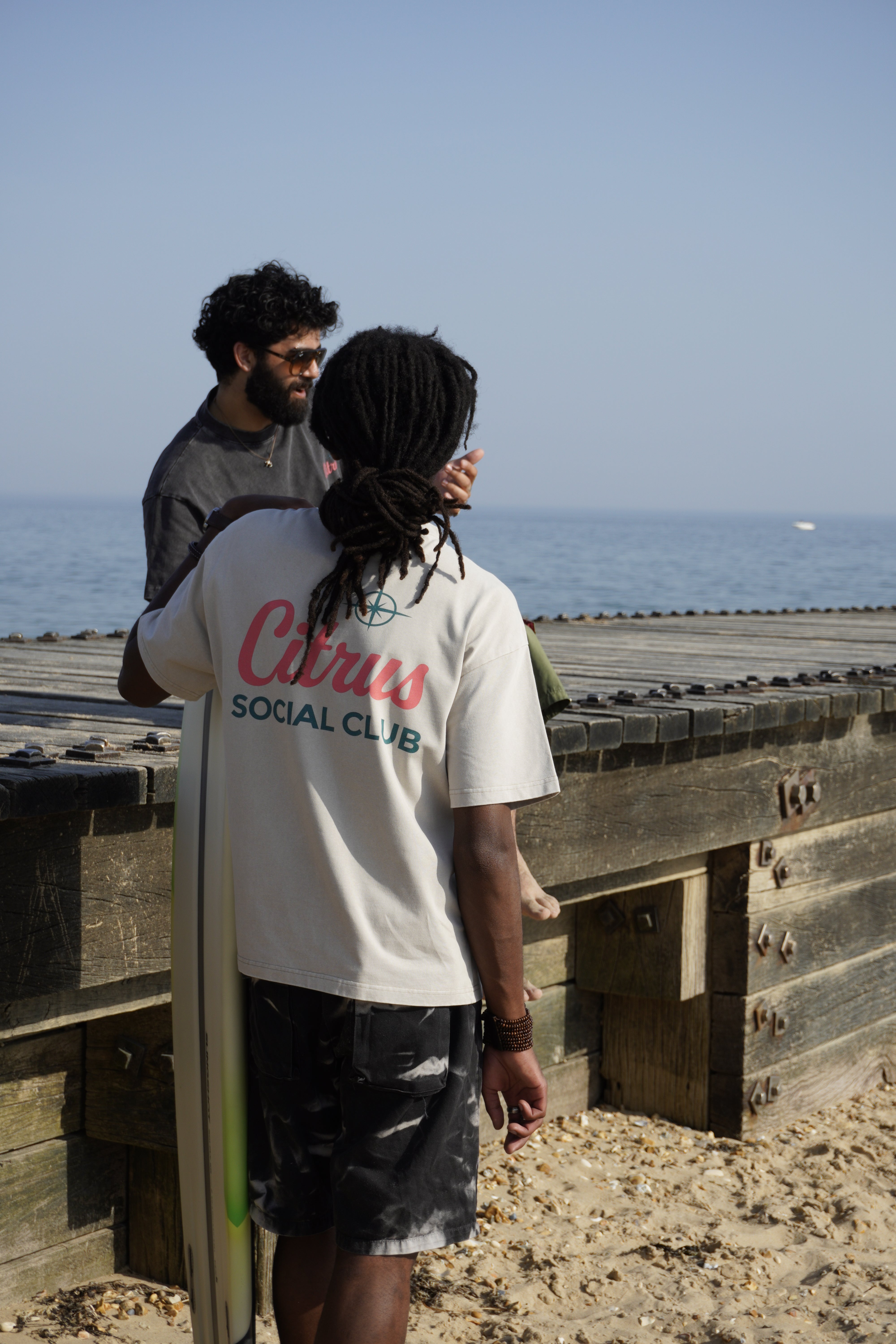 Two men standing on a wooden boardwalk by the ocean, with one holding a surfboard. The man in front wears a white t-shirt with 'Citrus Social Club' printed in red and green on the back, paired with black shorts.